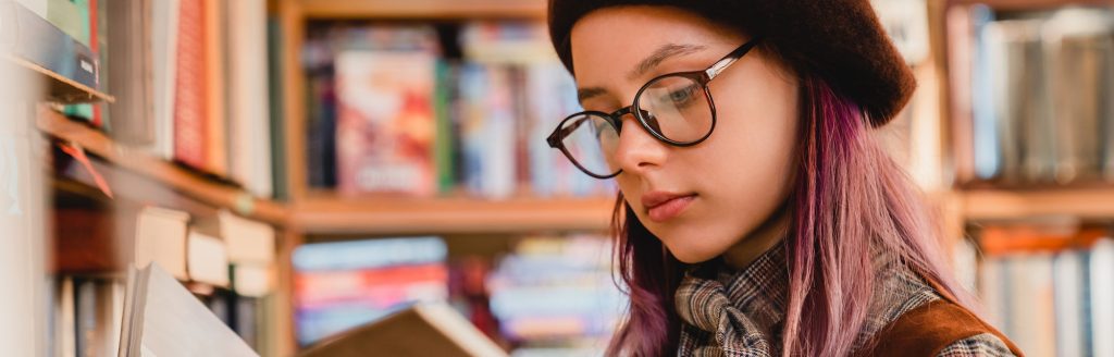 Intelligent young caucasian girl in glasses reading a book at the library